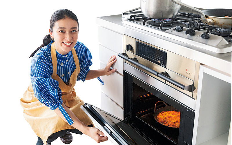 キッチンでオーブンの扉を開けるエプロン姿の武井咲さん
