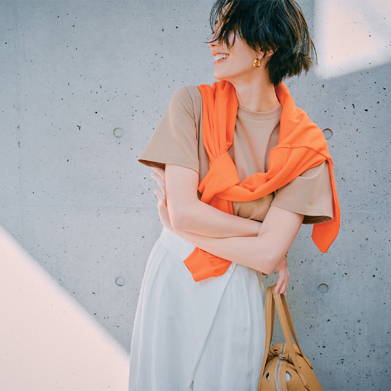 ベージュTシャツに白パンツを合わせアクセントにオレンジ色のニットを肩掛けしたコーディネート